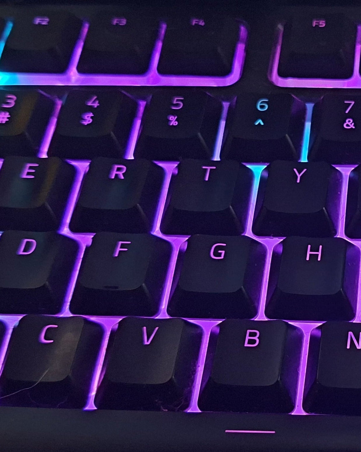 A close-up of a black mechanical keyboard with vibrant purple and blue RGB backlighting, photographed from a high angle.