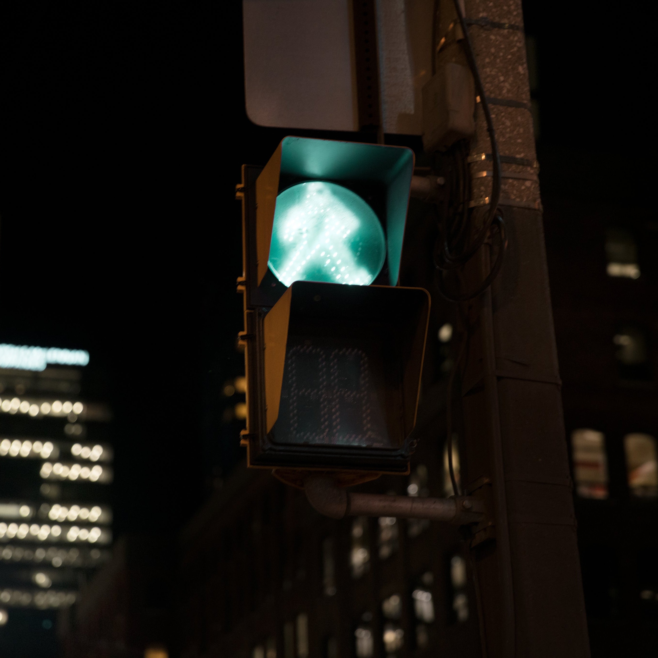 A street traffic signal at night showing a glowing green pedestrian walkway symbol, mounted on a pole with blurred city buildings in the background.